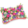 A pillow-shaped sympathy cushion photographed from a slightly elevated, oblique angle against a clean white background, its surface a sumptuous carpet of fresh blooms. The centre is dominated by velvety hot pink roses, some tightly budded and some gently unfurling, surrounded by pale shell-pink carnations that lend a soft, ruffled texture. Scattered through the composition are deep purple lisianthus and cheerful lemon-yellow ranunculus, their round, layered petals catching the light and adding lively contrast. Cool, architectural notes come from silver-blue Eryngium thistles placed like small stars among the petals, while sprays of silvery eucalyptus and mixed lush greens give depth, scent and a natural frame. The perimeter is neatly defined by a ruffled, deep red satin ribbon base - a pleated edge that echoes the cushion shape and adds a formal, tactile finish. The overall palette of fuchsia, violet, lemon and blush reads as a celebration of life rather than sombre mourning; you can almost imagine the faint sweet perfume of roses mingling with the resinous eucalyptus. Handcrafted with careful symmetry and structural support underneath, this floral pillow is intended as a comforting memorial piece for a funeral service or to rest gently at a graveside. Its presence softens a room with colour and texture, offering a visual embrace and a quiet message of remembrance from a local florist serving the Brent Cross area, mindful of nearby services and community moments of farewell.