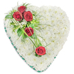 In this photograph, a traditional heart-shaped funeral arrangement is displayed against a bright white background, its form clear and uncompromised. The heart is completely covered with densely arranged white chrysanthemums, each small flower carefully placed so that together they form a luxuriously soft, cushion-like surface. The white petals give the impression of calm and purity, suggestive of peace and gentle rest. Arcing diagonally across the upper left area of the heart is a striking grouping of deep red roses, five in total. Three roses are clustered near the top left curve, their velvety petals opened just enough to show depth and richness, while two more sit slightly lower and to the right, maintaining a flowing line that leads the eye. Dark, glossy foliage, delicate fern-like sprigs, and tiny white filler flowers surround and support the roses, adding contrast and bringing the design to life with varied textures. Slender blades of bright green ornamental grass sweep across the heart in soft curves, visually linking the roses and suggesting a sense of continuity and enduring connection. Around the outer edge, a deep green satin ribbon is neatly pleated to frame the heart, creating a defined border that enhances the overall shape without distracting from the flowers. The careful craftsmanship and serene colour palette make this piece a fitting choice for memorials, funerals, or graveside visits in the Brent Cross area, whether delivered to a local chapel, crematorium, or family home. It conveys a message of love, respect, and remembrance, expressed through the quiet beauty of white chrysanthemums and the profound symbolism of red roses.