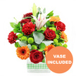 In this image, a colourful, upbeat bouquet is displayed in a modern rectangular glass vase, evoking the energy and warmth of a celebration in Brent Cross. The vase is crystal clear, showing the neatly gathered stems inside, and is wrapped around its lower portion with a light green and white gingham ribbon that introduces a gentle, homely charm. Positioned centrally are several deep red roses, lush and velvety, their tightly layered petals forming rich, romantic blooms that anchor the design. Mixed among them are ruffled red-orange carnations, each flower a mass of finely crimped petals that create soft texture and add an inviting, almost cloud-like fullness. Bright orange gerbera daisies with dark centres punctuate the arrangement, their flat petals radiating like sunbeams and bringing a sense of joy that calls to mind a bright afternoon near Brent Cross Shopping Centre. Elevated above the main cluster are white Oriental lilies, some already open into star-shaped blossoms with gentle pink veining along the inner petals, others resting in smooth, unopened buds, promising continued unfolding beauty over the coming days. Scattered throughout are small, perfectly spherical lime-green pompom chrysanthemums, their fresh, vivid colour adding a cool counterpoint to the warm reds and oranges while enhancing the sense of abundance. Surrounding the blooms is a generous frame of greenery: broad, glossy dark green leaves and soft, silver-green eucalyptus sprigs layer over one another, giving depth, movement, and a natural garden feel, as though arranged moments ago by a skilled local florist for delivery around the North Circular. The entire composition sits against a stark white background, which intensifies each hue and detail. In the lower right corner, slightly overlapping the bouquet, an orange circular badge with crisp white capital letters reads VASE INCLUDED, clearly indicating that the stylish glass vase forms part of this ready-to-display floral gift.