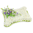 A comforting, rectangular sympathy pillow crafted entirely from hundreds of small, pristine white chrysanthemum heads sits serenely against a clean white backdrop. The chrysanthemums are densely packed to form a plush, pillow-like surface - a soft, even field of snowy petals that reads at once as tender and resolute, suggesting quiet rest. The pillow's edges are defined by a delicate, light green satin ribbon that has been pleated into a gentle scalloped trim; this fresh, lime-toned border lifts the composition with a whisper of renewal and gives the piece a refined finished edge. At the upper left the arrangement carries a restrained, elegant spray of accent flowers: layered white lisianthus mingle with deeper purple lisianthus blooms and a few tightly furled purple buds, their velvety tones creating a thoughtful contrast against the white bed below. Intertwined fern fronds and graceful green foliage add filigree texture and a living, airy quality, while a broad dark-green leaf wrap secures the stems and keeps the accent neat and composed. Soft studio lighting creates a faint drop shadow beneath the pillow, highlighting its slightly raised, succulent profile and the artisan's careful shaping. The overall scent is faintly floral and clean - the gentle green notes of foliage balancing the mild, powdery chrysanthemum perfume. Intended for memorial services, graveside placement, or a quiet viewing, this tribute evokes peace, dignity, and consolation. An experienced local florist might recommend it for wakes or chapel services in the Brent Cross area, from nearby Cricklewood to the precincts around the shopping centre, where its subdued beauty offers comfort without overwhelm.