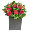 A composed, modern bouquet of rich crimson roses sits like a small sculpture; each bloom opens in velvety spirals, the petals deep and saturated with romantic red, their centers drawing the eye inward. Around the roses, broad glossy leaves have been artfully curled and layered to create a rhythmic frame - overlapping emerald waves that catch the light and give the composition a sense of movement and depth. The arrangement rests in a matte black square vase whose clean edges and understated finish lend a contemporary, urban confidence to the piece; the dark vessel visually anchors the vibrancy above and sets off the crimson like a jewel. Studio lighting is bright and clear, skimming the surface of each petal and flashing highlights along the waxy foliage, so the picture feels crisp and the textures read distinctly: silky rose petals, firm structured leaves, and the tactile matte of the container. Close to the blooms there is the imagined scent of fresh rose, softened by green, almost lemony top notes from the leaves - a subtle perfume that lifts the classic sweetness into something more modern and fresh. As a local florist mindful of place, I picture this arrangement bringing warmth to a Brent Cross sitting room or making a confident statement on a table near Claremont Park or Cricklewood Broadway. It is crafted with intention: suited to anniversaries that call for dramatic affection, to thank-you moments that demand elegance, or to urban interiors that need a striking, quietly passionate focal point. The overall effect is refined, tactile, and emotionally resonant, a bouquet that reads both as art and as heartfelt message.