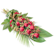 A long, gently tapered spray of red roses and foliage is displayed diagonally across a crisp white background, its poised composition reflecting the thoughtful care often given to funeral tributes in the Brent Cross community. The arrangement begins at the top left where the stems are neatly aligned and closely bound with a broad, folded green leaf wrap, giving the piece a tidy and natural finishing touch. From this point, the design opens out into a flowing succession of red roses, each bloom a rich, velvety crimson. Some roses remain in bud, their petals wrapped tightly in promise; others are partway open, revealing spiralled centres that catch soft highlights. Nestled between the roses, clusters of small, round Hypericum berries in a pinkish-red shade add detailed texture and a slightly brighter note, lightening the depth of the overall red palette. A strong foundation of dark green foliage supports the flowers, with broad, glossy leaves overlapping to form a continuous bed of green that feels both structured and comforting. From the lower right, graceful palm fronds extend in fine, feathery lines, stretching the arrangement into an elegant, elongated profile and adding a sense of quiet movement, almost like a gentle bow. Against the pure white backdrop, the contrast of vivid reds and deep greens becomes especially striking, while still maintaining a sense of calm and reverence. This classic red rose funeral spray would sit beautifully at a service near Brent Cross Shopping Centre, in a local church along Hendon Way, or by a graveside, offering a simple, honest expression of love, remembrance, and respect for someone dearly missed.