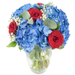 A bright, tactile bouquet sits in a simple clear glass vase against a clean white backdrop: large, cloud-like clusters of sky-blue hydrangea form the generous, rounded core, each tiny floret overlapping like raindropped petals and cooling the eye with a soft cobalt glow. Nestled through that blue volume are three deeply saturated red roses, their velvety petals still tightly spiralled at the centre, punctuating the composition with a confident, romantic note. Airy sprays of ivory baby's breath drift between the larger shapes, lending a delicate, star-like lightness that lifts the arrangement, while rounded, silvery-green eucalyptus leaves weave through the stems and introduce a muted herbal scent and soft, matte texture. The glass vase narrows gently at the neck and reveals the fresh green stems submerged in clear water, a reassuring sign of freshness, and the whole arrangement is lit from above in bright, natural light so every curve and shadow reads clearly. As a local florist would note, the palette of cool blue, passionate red and pure white feels both celebratory and quietly refined - equally at home as a centrepiece on a Brent Cross dining table, a thoughtful anniversary gift delivered to a Clitterhouse Road flat, or a patriotic nod at a small community gathering near the Shopping Centre. The bouquet balances plush volume with airy detail; you can almost sense the soft, leafy fragrance of eucalyptus mixed with the faint, sweet perfume of rose. Expertly composed to feel generous yet modern, this arrangement invites touch, scent and the small pleasure of bringing colour into a bright interior.