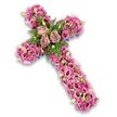 An intricate floral cross tribute, bathed in soft pink, mauve, and lilac tones, is photographed against a bright white background, its fine details brought into sharp focus. The cross's vertical stem and outstretched arms are fully covered in a thick layer of lisianthus blooms, whose delicate, slightly ruffled petals interlock to create a velvety, cushion-like surface. The colours drift from pale blush and pastel rose along the edges to richer pinks and muted lavender tones closer to the centre, with select flowers showing darker, more intense cores that lend depth and a gentle sense of shadow. At the heart of the cross, where the two arms meet, three fully open roses in a dusty rose or light mauve shade form a calm, central point. Their petals are wide and layered, curling slightly at the edges as if caught at the peak of their bloom. Surrounding this trio, feathery fern fronds and compact, glossy green leaves create pockets of contrast and texture, adding a cool freshness to the warm pinks. Tiny pink and purple accent blossoms, together with small cream and white flowers holding golden-yellow centres and a scattering of pale green buds, weave through the main flowers, giving the arrangement a handcrafted, detailed feel. Along the perimeter of the cross, slight glimpses of deep purple ribbon or structural backing appear now and then, subtly outlining the traditional shape. The piece exudes quiet dignity and comfort, much like a floral cross carefully arranged by Brent Cross Flowers for a local church or crematorium service, offering a touching expression of faith, memory, and gentle support for grieving families.