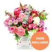 In this image, a sumptuous mixed bouquet brimming with roses, carnations, and daisy-style blooms is presented in a clear square glass vase set against a spotless white background. The central focus is on the roses, which range from intense magenta and fuchsia shades to gentle, romantic blush pinks, their velvety petals arranged in spirals that invite a closer look. Tucked closely around them are lush carnations in coral, peach-pink, and creamy white, their characteristic ruffled petals adding body, softness, and a sense of movement to the design. Interwoven among the larger flowers are smaller lavender and purple chrysanthemums with vivid green centres, providing little bursts of cheerful contrast that echo the lively spirit of the Brent Cross area. A generous collar of greenery encircles the blooms: rounded eucalyptus leaves with a soft, silvery-green tone, darker green foliage for structure, and a few hints of reddish-brown leaves that lend a warm, slightly autumnal nuance. The stems are neatly aligned inside the clear vase, which is trimmed at the base with a white band decorated with repeating red heart icons and the handwritten-style phrase all my love, turning the bouquet into a clear declaration of affection. In the bottom right corner, an orange circular badge with clean white capital letters reading VASE INCLUDED stands out, indicating that the flowers arrive already arranged and ready for immediate display on a table or desk. The overall impression is of a carefully composed, ready-to-gift floral arrangement from a trusted Brent Cross florist, ideal for deliveries to nearby Hendon, Dollis Hill, and surrounding streets when celebrating birthdays, anniversaries, or simply sending warm thoughts and love.