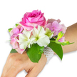 A refined wrist corsage rests against a clean white background, shown on a left hand and forearm with a light-to-medium skin tone so the florals read clearly against skin. The arrangement centres on a large, fully opened deep fuchsia-pink rose whose layered petals curl inward and outward, catching a soft, diffuse light that suggests a gentle studio glow. Around the rose, clusters of blush pink hydrangea florets create a plush, cloud-like base: their papery, slightly ruffled edges add both volume and an airy softness. Crisp white alstroemeria (Peruvian lilies) fan out in small sprays, each petal bearing a faint pale green streak toward the throat that draws the eye to the rose. On one side a tight cluster of light green alstroemeria buds and a few tiny, barely open pink buds promise more blooms, their satiny surfaces contrasting with the hydrangea's matte texture. Broad, glossy emerald green leaves cradle the composition, their rich colour grounding the pastels and lending a visual weight that feels both fresh and sophisticated. The whole is secured to the wrist by a delicate, multi-strand bracelet of small lustrous white pearls that wrap snugly and trail slightly across the back of the hand, glimmering against skin like quiet jewellery. The scent, if imagined, would be a soft, powdery rose note over green undertones. This wearable bouquet, with careful wiring and tape hidden beneath the foliage, reads as a polished choice for prom, a bridesmaid's accessory, or an intimate celebration where classic elegance and tender craftsmanship are required.